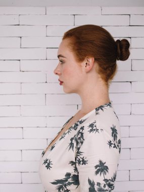 Beyaz tuğla duvarın önünde duran, güzel elbiseli, çekici, düşünceli, kızıl saçlı kadının profili.