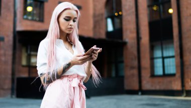 Uzun düz, parlak gül saçlı, yaz kıyafetleri giymiş, caddede beklerken cep telefonuna göz atan genç ve modaya uygun bir kadın.