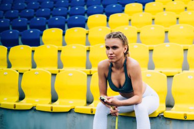 Sarı örgülü spor kıyafetli bayan antrenör boş mavi ve sarı stadyum tribünleri bekliyor.