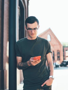 Şık giyinmiş ve gözlüklü konsantre erkek öğrenci şehir caddesinde arkadaşlarıyla mesajlaşırken yığına yaslanıyor.