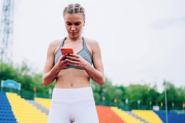 Spor giyim sektöründe kendine güvenen ve güzel bir bayan stadyumda elinde akıllı telefonuyla duruyor ve sosyal medyada bulanık arka planda mesajlaşıyor.
