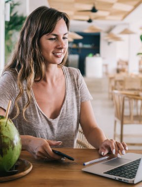 Genç milenyum kadını hindistan cevizi kokteyli ile masada oturan dizüstü bilgisayarın ayarlarını kontrol ediyor ve internet iletişimi için kafeterya web bağlantısı kullanıyor, dijital göçebe yaşam tarzı kavramı