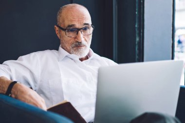 Beyaz tişörtlü, siyah pantolonlu, dizüstü bilgisayarlı yaşlı, sakallı adam oturma odasında pencerenin yanında oturuyor.