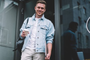 Kot ceketli, elinde bir fincan kahve ve dizüstü bilgisayarla caddede kameraya gülümseyen mutlu genç adamdan.