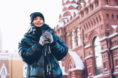 Koyu renk kışlık ceketli tatmin olmuş genç bir kadın açık gökyüzünde plastik bardakta sıcak kahveyle tarihi binaların yanında duruyor.