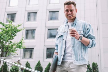 Sokağın ortasında kameraya gülümseyen, tek kullanımlık kahve ve dizüstü bilgisayarlı yakışıklı genç adamdan.