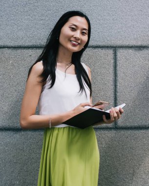 Rahat stil sahibi genç Asyalı kadın planlayıcısı telefonda gezerken şehir merkezindeki ofis binasının yanında gündüz vakti dikiliyor.