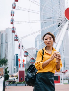 Hong Kong caddesinde dönme dolaba gülümserken, elinde kahve fincanı tutan sıradan giyinmiş, mutlu, genç bir kadın.