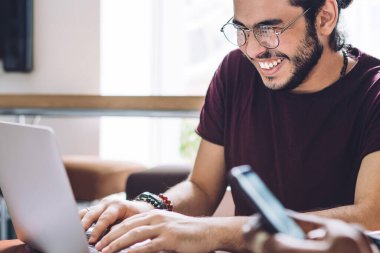Gözlüklü neşeli bir adam dizüstü bilgisayarını kullanıyor. Not defteriyle oturuyor ve grup arkadaşlarıyla gülüyor.