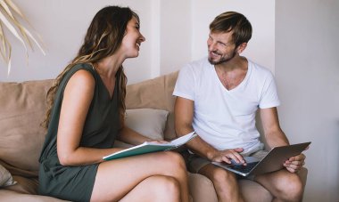 Diz üstü bilgisayarı karıştıran neşeli yakışıklı bir adam ve elinde not defteri olan mutlu bir kadın evdeki kanepede oturmuş birbirine bakıyor.