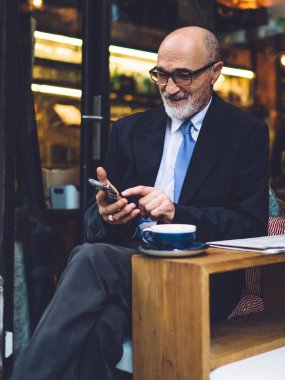 Gözlüklü ve takım elbiseli yaşlı, gülümseyen bir adam sokakta akıllı telefon kullanırken bacak bacak bacak üstüne atmış oturuyor.