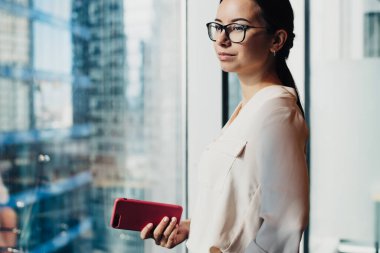 Gözlüklü genç, kendine güvenen iş kadınının elinde kırmızı bir akıllı telefon tutarken ve pencerenin önünde dikilirken düşünceli bakışları.