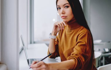Ciddi esmer kadın çalışanın portresi dizüstü bilgisayarla oturmuş araştırma ve muhasebe yapıyor, çekici kendine güvenen iş kadını kameraya bakıyor ve pencerenin yanında notlar alıyor ve planlar yapıyor.
