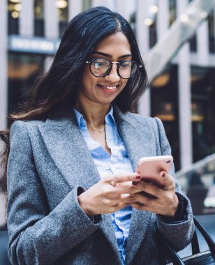 Gözlüklü ve modaya uygun ceketli modern iş kadını New York şehrinin arka planında cep telefonu kullanarak duruyor.