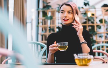Şık, mutlu, pembe saçlı, günlük kıyafetleri olan, akıllı telefondan konuşan ve hipster kafeteryasında oturmuş çay içen bir kadın.