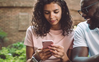 Gözlüklü, gülümseyen hippi, akıllı telefon ekranına bakarak sevgili kız arkadaşının yazdığı SMS mesajlarını kontrol ediyor. Aşık çift akıllı telefon cihazlarında 4G kablosuz internet kullanıyor.