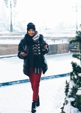 Kağıt bardak ve telefonla kışın gülümseyen genç bayan karlı caddede Noel ağaçlarının yanında yürüyor ve kameraya bakıyor.