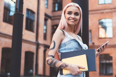 Not defterleriyle gülümseyen ve başını çeviren neşeli genç bir kadının yan görüntüsü. Şehir caddesinde kolona yaslanıp akıllı telefon kullanıyor.