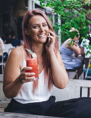 Elinde smoothie ile kafedeki beyaz ahşap bankta oturan telefonda kameraya bakarak konuşan güzel, neşeli bir kadın.