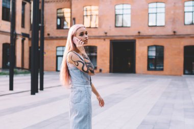 Uzun pembe saçlı, elinde dövmesi olan, büyük bir eğitim merkezinin iç bahçesinde cep telefonuyla konuşan kadının yan görüntüsü.