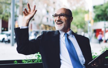 Zevkli gri sakallı, gözlüklü ve mavi kravatlı yaşlı adam kafenin önünde oturmuş tablet kullanırken el sallıyor.