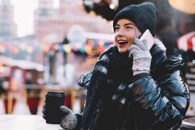 Kışlık ceketli iyimser modern kadın akıllı telefon kullanıyor ve bulanık arka planda süslenmiş Noel meydanında kahve içiyor ve yüzünü çeviriyor.