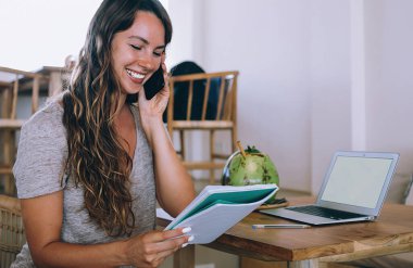 İyimser tatmin olmuş, rahat bir sandalyede oturan, kağıt okuyan, dizüstü bilgisayar kullanan ve kafeteryada akıllı telefondan konuşan bir kadın.