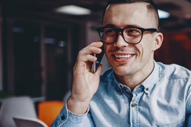Gözlüklü, gözlüklü, akıllı telefonlu, arkadaşlarıyla iletişim kuran, modern çalışma ortamında masada oturan neşeli, kot gömlekli, cep telefonuyla gülen genç bir adam.