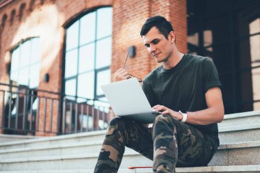 Ekrana odaklanan gözlüklü, askeri tarzda giyinmiş ve merdivenlerde otururken dizüstü bilgisayar kullanan düşünceli bir adamdan.