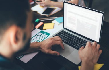 Bulanık ekin adam modern dizüstü bilgisayarda çalışıyor ve arka planda kağıtlar, malzemeler ve iş arkadaşıyla masada otururken daktilo yazıyor.