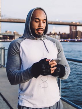 Genç kaslı Afrikalı Amerikalı adam ayakta durup kulaklıkla müzik dinlerken nehir ve köprünün arka planına akıllı telefondan mesaj atıyor.