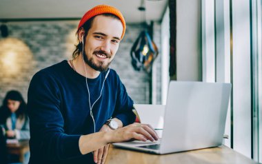 Mutlu gülümseyen hipster adamın portresi kafeteryada öğrenim görürken favori müzik listesi için zaman harcıyor, kulaklıklı pozitif bir genç evde dizüstü bilgisayarla masada oturuyor.