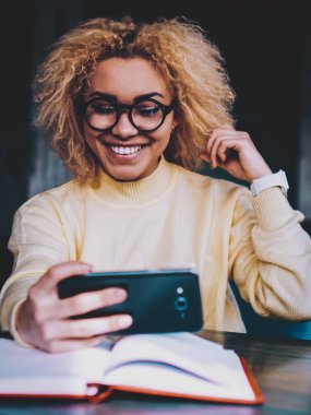 Görsel düzeltme için şık gözlüklü mutlu Afro-Amerikan bayan gençlik. Bilgi ve otomasyon için kafeteryada geçirilen zamanda cep telefonu cihazıyla selfie fotoğraflarına tıklayarak.