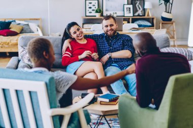 Neşeli bir grup erkek ve kadın hippi oturma odasında keyifli bir şekilde sohbet ediyorlar. Kafkasyalı mutlu bir çift Afro-Amerikan misafirlerle konuşuyorlar. Şık bir dairede koltukta oturuyorlar.