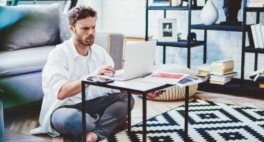 Ciddi bir beyaz erkek, dizüstü bilgisayarla online rezervasyon yapıyor modern iç oturma odasında oturuyor, serbest çalışan adam evde netbook kullanarak uzaktan çalışmaya odaklanmış.