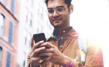 Gözlüklü, kahverengi süet ceket giyen, New York caddesinde güneşli bir günde gülümseyen ve cep telefonuyla mesajlaşan yakışıklı, tıraşsız İspanyol erkekten.