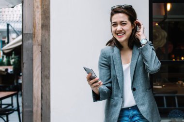 Güzel, güzel, mutlu, resmi giyinmiş, akıllı telefon kullanan, duvara yaslanmış, caddede kameraya bakan bir kadın.