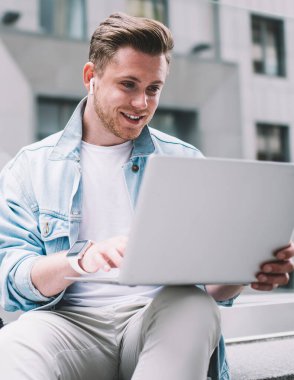 Günümüz kıyafetleri ve kulaklıkları olan, çantalı ve kahveli modern yakışıklı adamı dizüstü bilgisayarla çalışıyor.