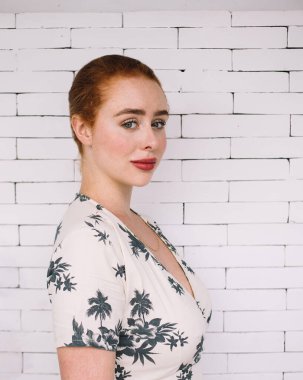 Beyaz tuğla duvarın önünde dikilip kameraya bakan yaz elbiseli, güler yüzlü kızıl saçlı kadının yan görüntüsü.