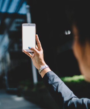 Siyah ceketli genç bir kadın güneşli bir günde şehir merkezinde cep telefonuyla fotoğraf çekiyor.