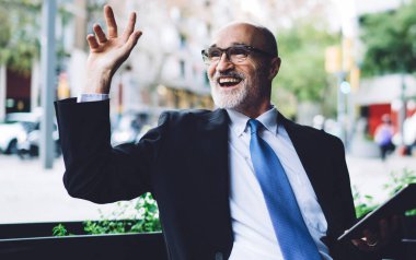 Zevkli gri sakallı, gözlüklü ve mavi kravatlı yaşlı adam kafenin önünde oturmuş tablet kullanırken el sallıyor.