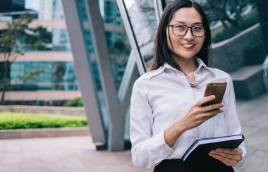 Gözlüklü güzel bir Asyalı kadın ve ellerinde defteriyle gülümsüyor ve akıllı telefon kullanıyor. Şehir caddesinde duruyor. Ofis binasının yanındaki kameraya bakıyor.