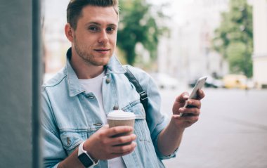 Kot ceketli, tek kullanımlık kahve içen ve sokakta duvara yaslanmış cep telefonu kullanan rahat bir adam.