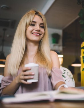 Alçak açılı, gülümseyen, açık pembe elbiseli, dizüstü bilgisayarda çalışan ve kahve dükkanının masasında bir fincan kahve tutan sarışın.