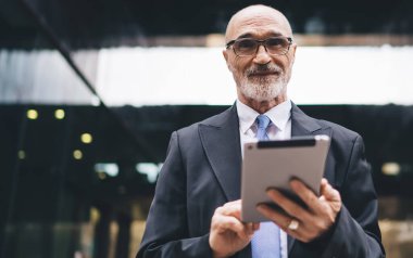 Yakışıklı, sakallı, kel, resmi kıyafetli ve gözlüklü bir adam iş merkezinin önünde kameraya bakıyor.