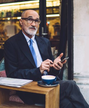 Gözlüklü, takım elbiseli, akıllı telefonlu yaşlı bir adam cama yaslanmış ve kahve fincanıyla yan yana oturuyor.