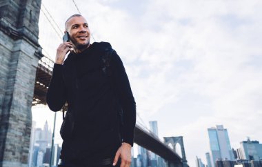 Gülümseyen, kısa saçlı, siyah giysili genç adam şehir planında güpegündüz köprüde akıllı telefonla iletişim kuruyor.