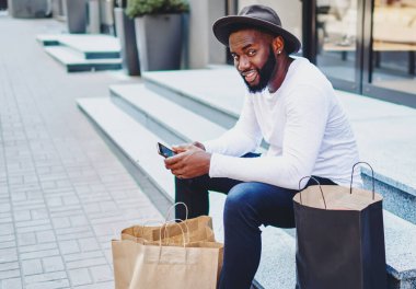 Ağ mağazası servisinden geribildirim bekleyen Afro-Amerikalı bir adamın portresi merdivenlerde oturmuş akıllı telefondan alışveriş sonrası dinlenen şık şapkalı, pozitif esmer bir adam.