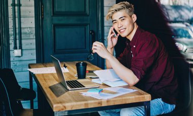 Akıllı telefon cihazını ararken kameraya gülümseyen Çinli pozitif hipster öğrencinin portresi. Dizüstü bilgisayarın başında oturan ve hücresel cihazla konuşan neşeli Japon genç adam.