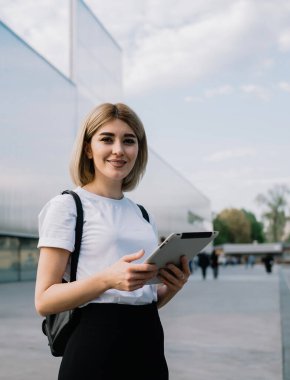 Günlük kıyafetlerle gezen neşeli kadın, şehir caddesinde ofis binasının yanında durup kameraya gülümserken zevkle tableti karıştırıyor.